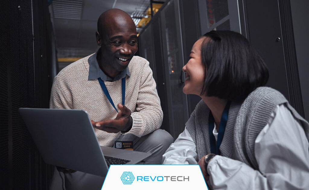 Two smiling IT professionals crouched by hardware and an open laptop discussing risk reduction.