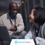 Two smiling IT professionals crouched by hardware and an open laptop discussing risk reduction.