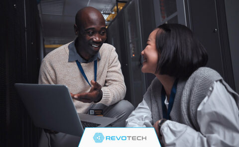 Two smiling IT professionals crouched by hardware and an open laptop discussing risk reduction.