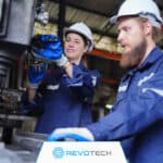 Image of two manufacturing workers in white hard hats and blue coveralls operating a machine at a manufacturing plant.
