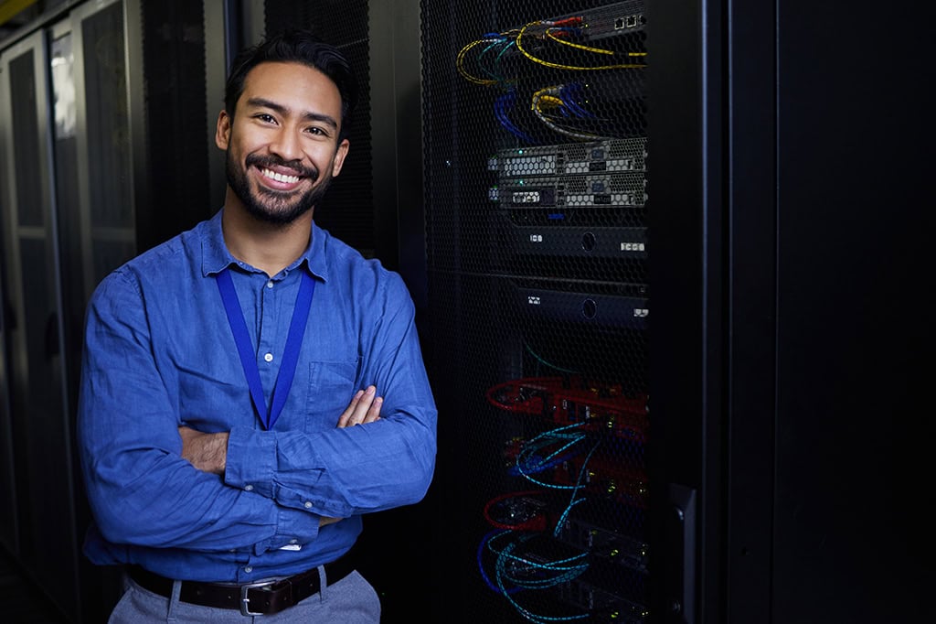 An IT support specialist stands next to a server farm smiling. An IT support specialist stands next to a server farm smiling.